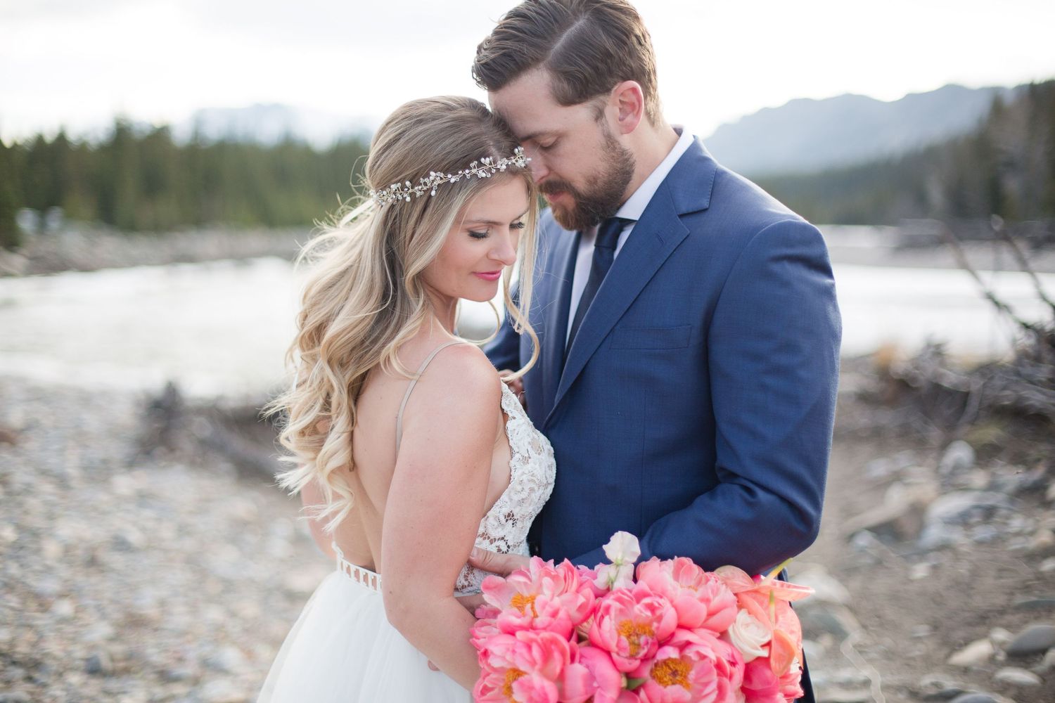 Couple in formal attire share intimate moment with pink bouquet in mountain setting.