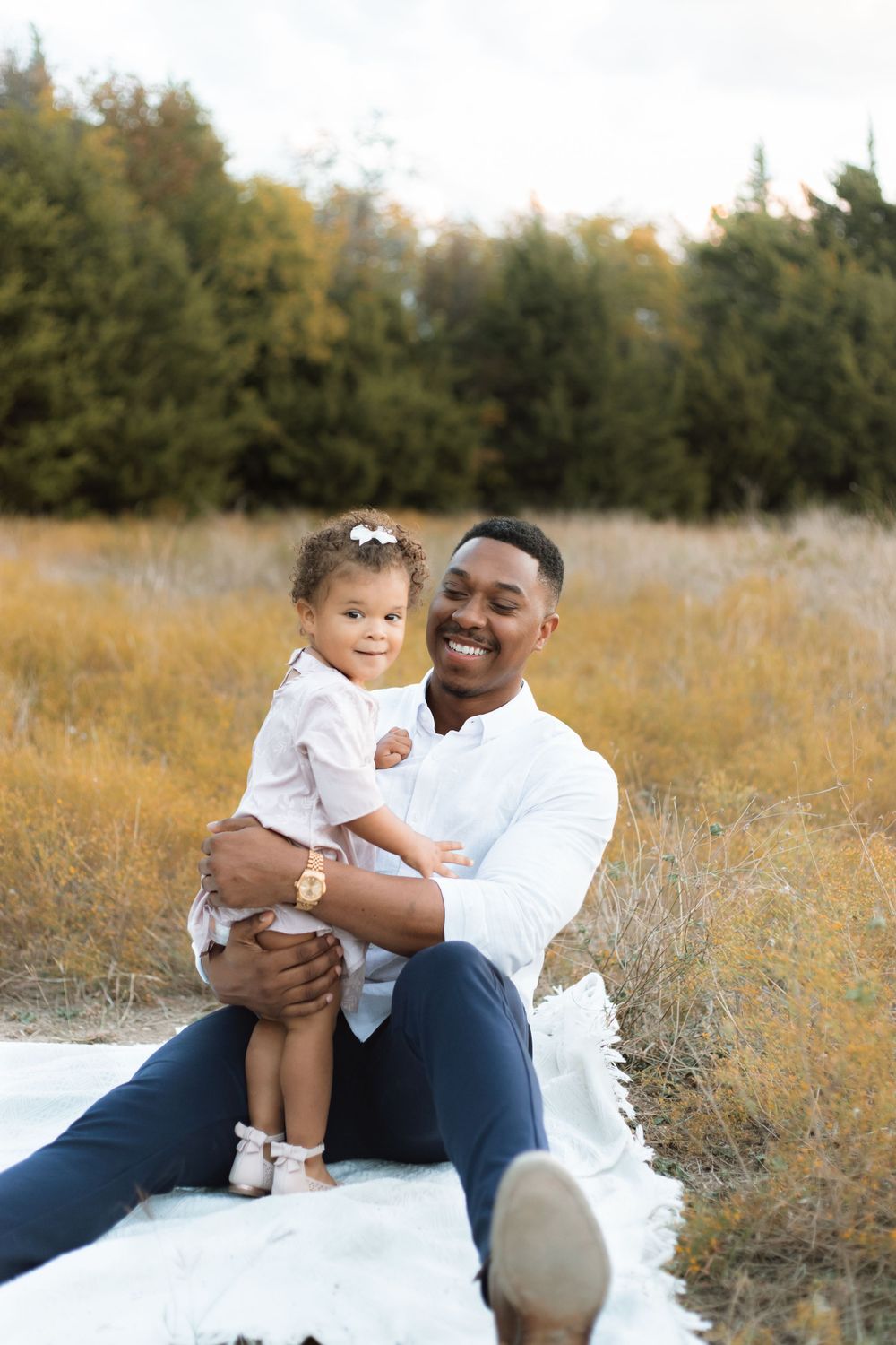 Casual outdoor family portrait with adult and toddler sitting on white blanket.