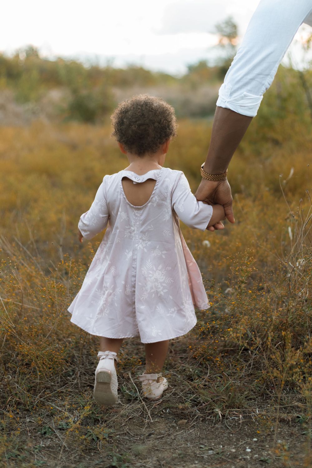 A toddler in a white dress holding hands with an adult while walking through a golden field.