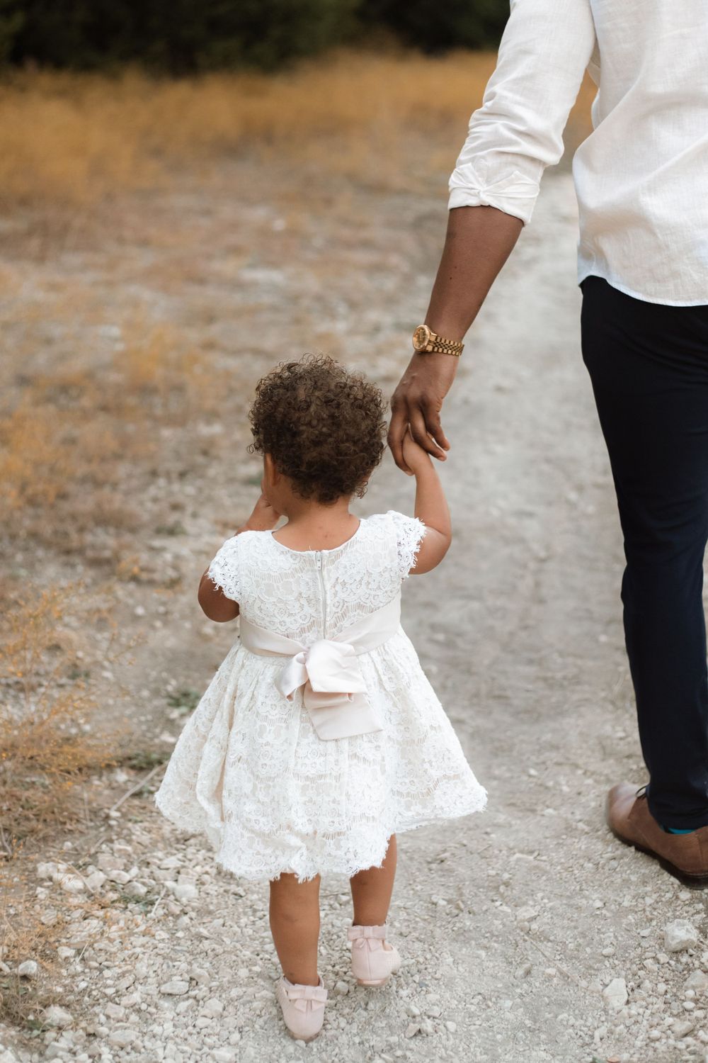 A small child in a white lace dress holds hands with an adult while walking on a path.