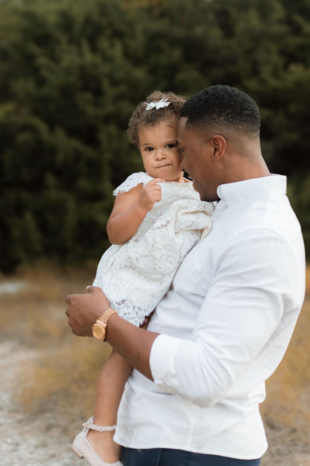 A tender outdoor portrait of an adult in white holding a small child wearing a lacy white dress.