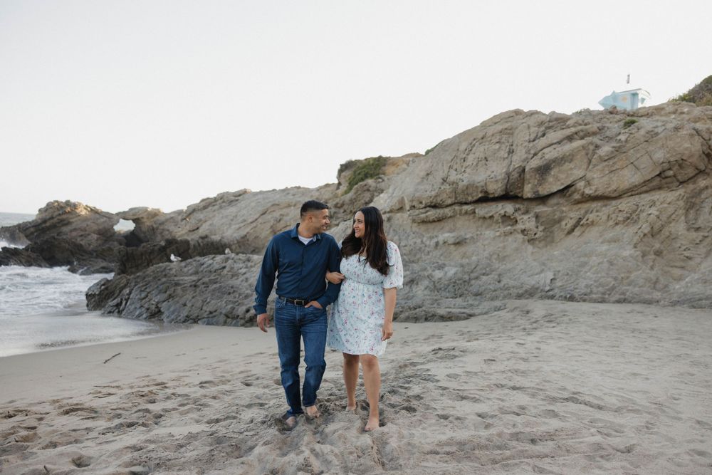 A romantic couple walking barefoot on sandy beach by rugged cliffs at sunset.