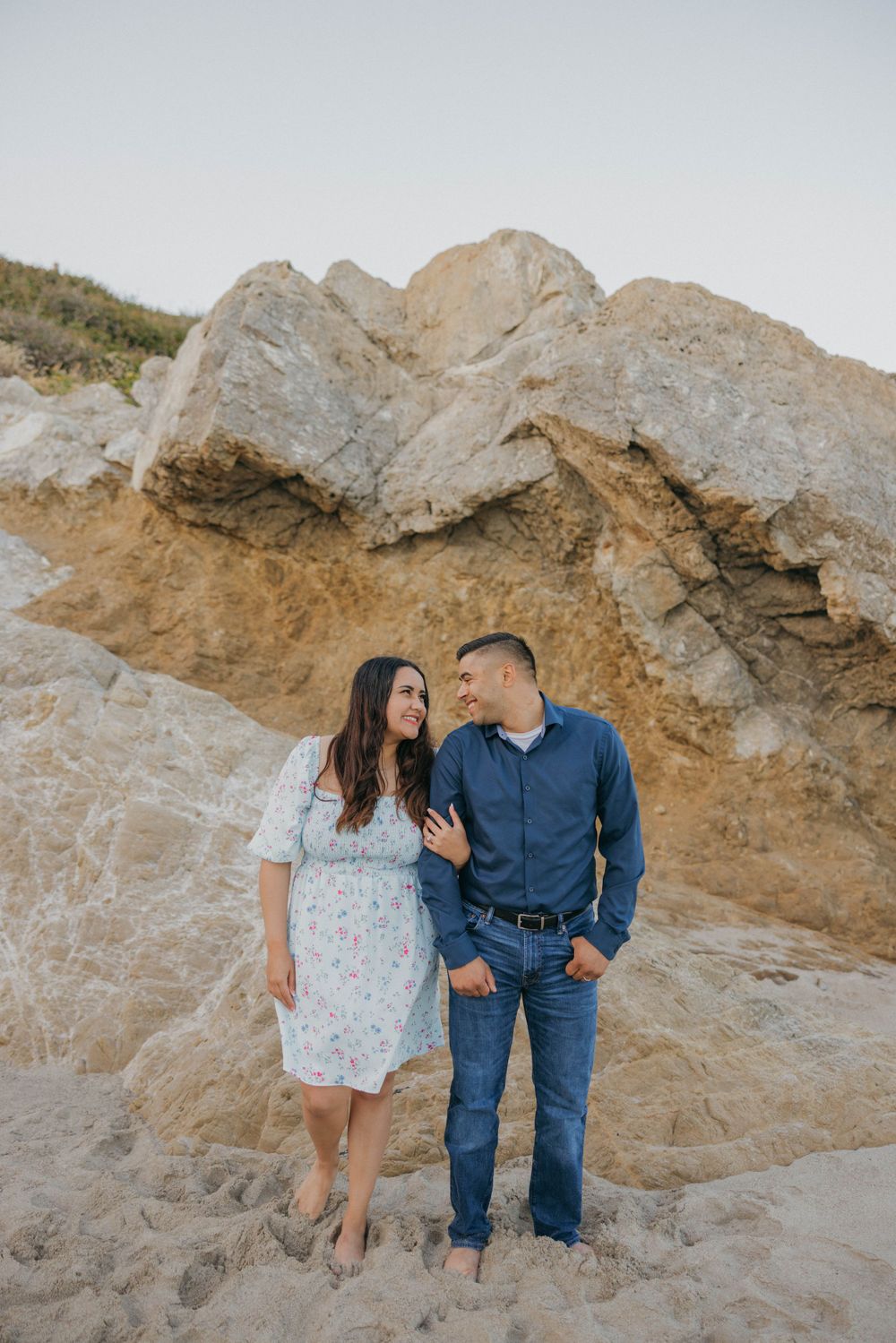 A couple standing close together against a dramatic rocky cliff backdrop at the beach.