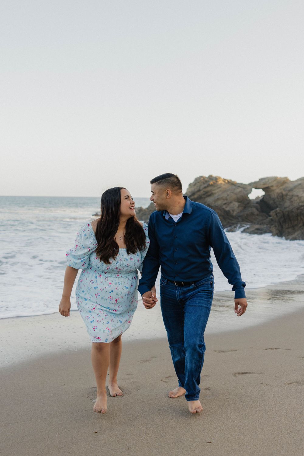 A couple walking barefoot on the beach wearing coordinating blue and white outfits.