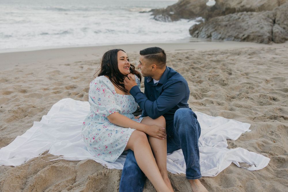 A romantic moment shared between a couple sitting on a white blanket at the beach.
