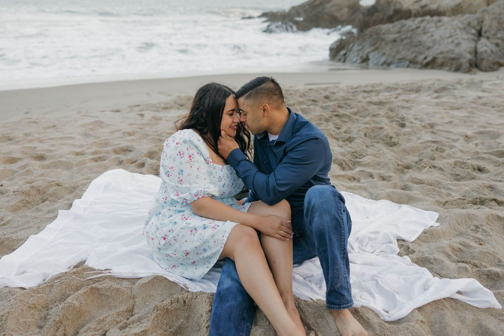 An intimate couple sharing a tender moment while sitting on a white blanket by the ocean.