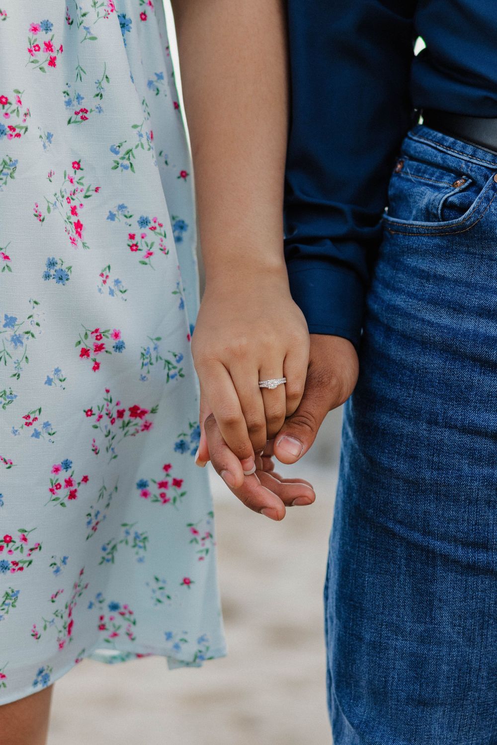A close-up of hands holding with an engagement ring visible against a floral dress and jeans.