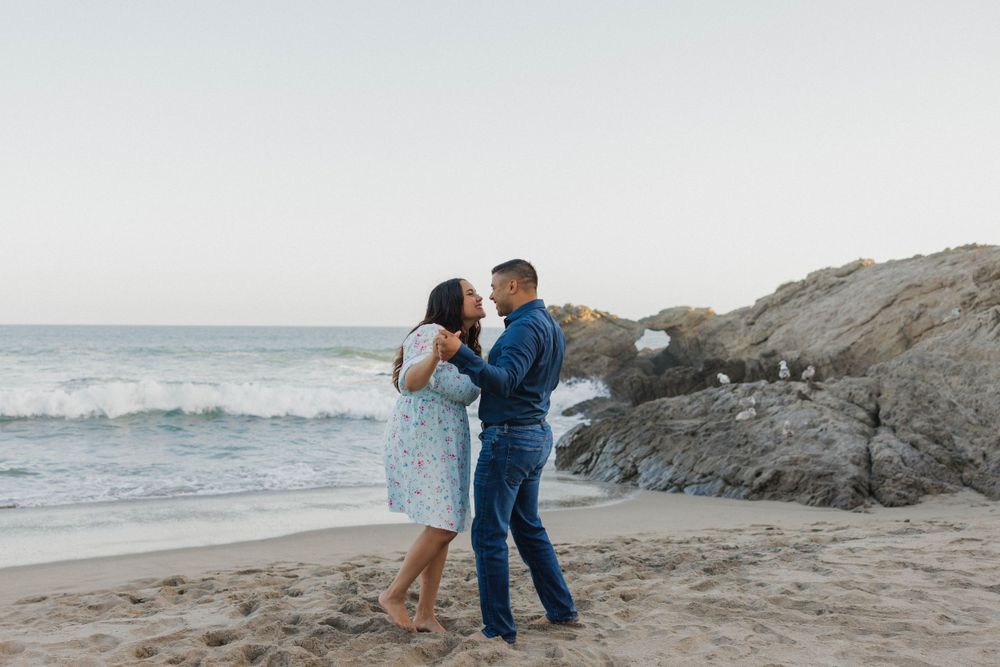 A romantic moment shared between a couple dancing together on a scenic beach with rocky cliffs at sunset.