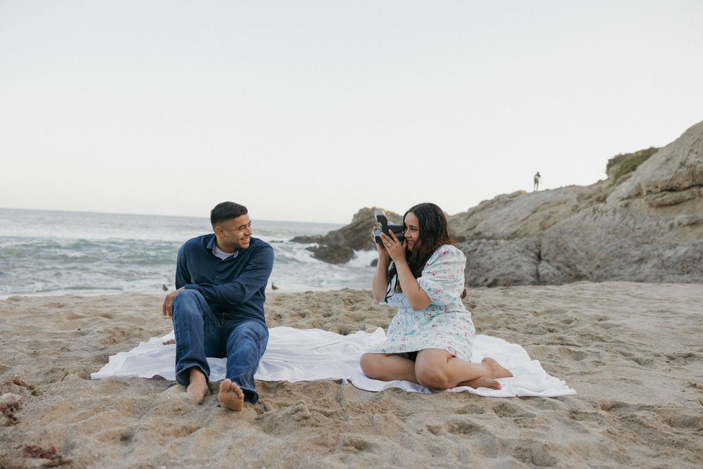 A playful photoshoot scene on a beach blanket with rocky coastal formations in the background.