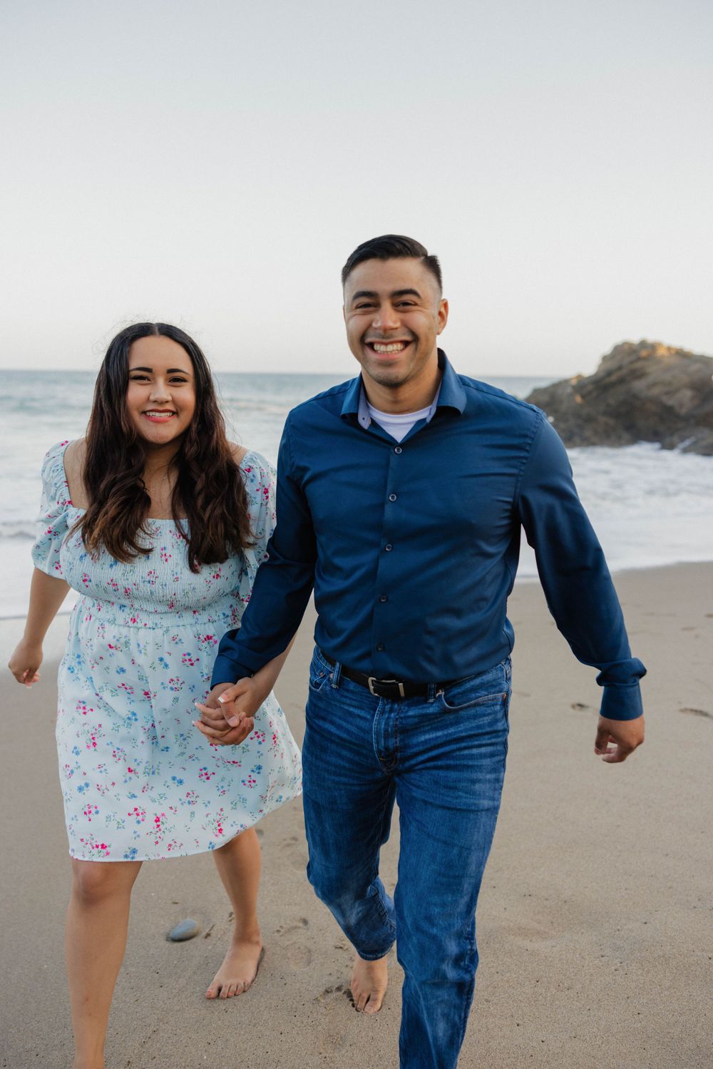 Joyful couple running barefoot along the shoreline wearing coordinated blue and floral outfits.
