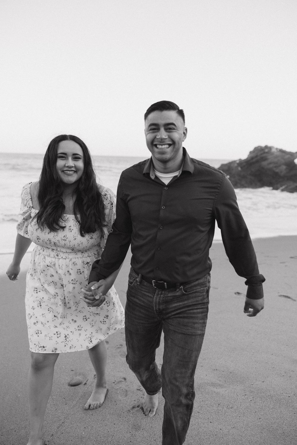 Happy couple walking barefoot on the beach in casual attire with rocky cliffs behind them.