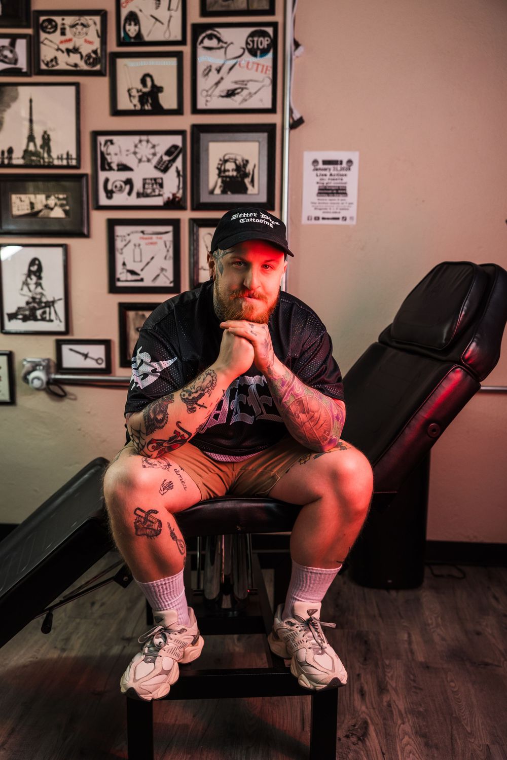 Heavily tattooed man in black cap sits in tattoo parlor chair surrounded by framed artwork walls.