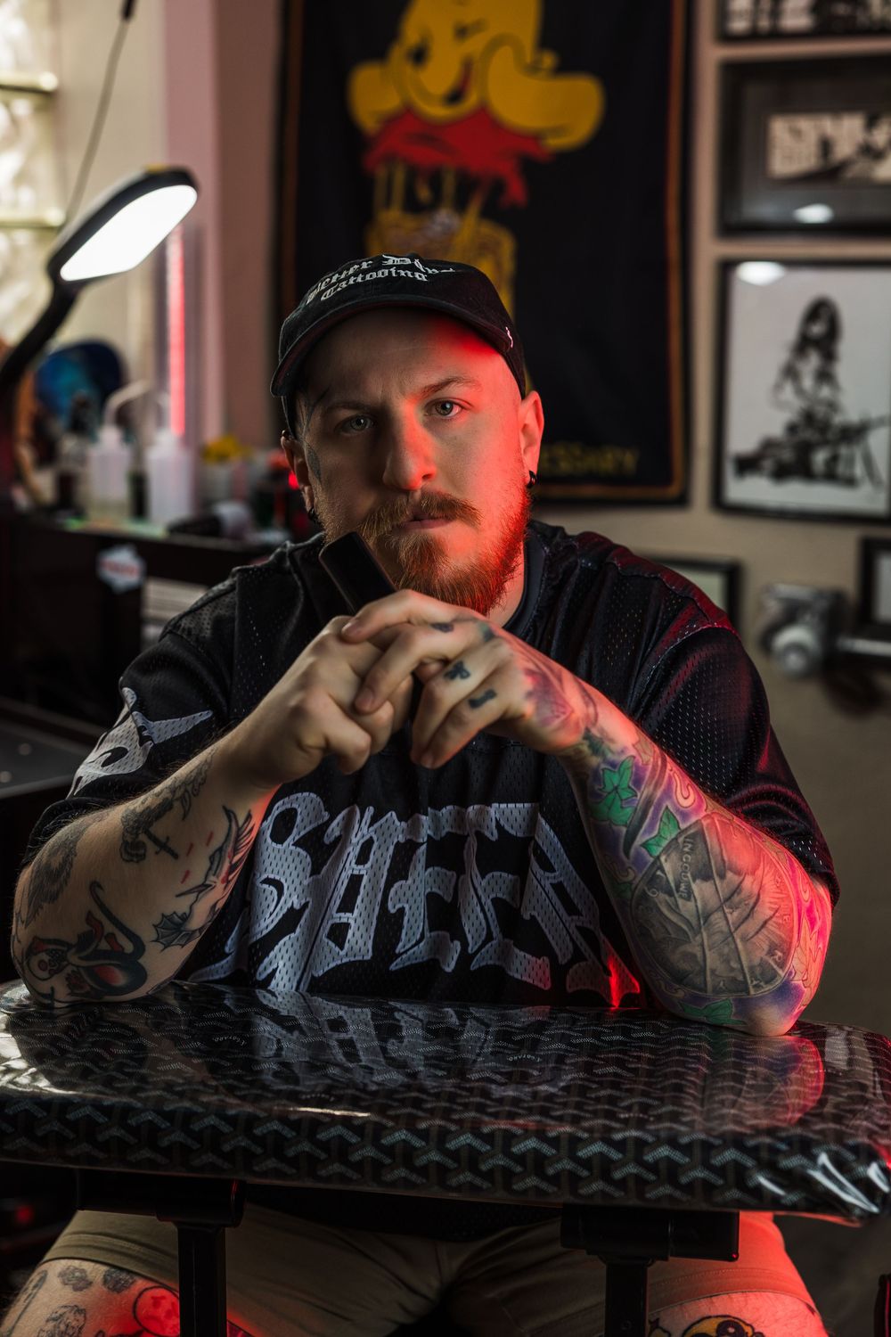 Tattooed bearded man in black graphic tee sits at tattoo studio table with hands clasped.