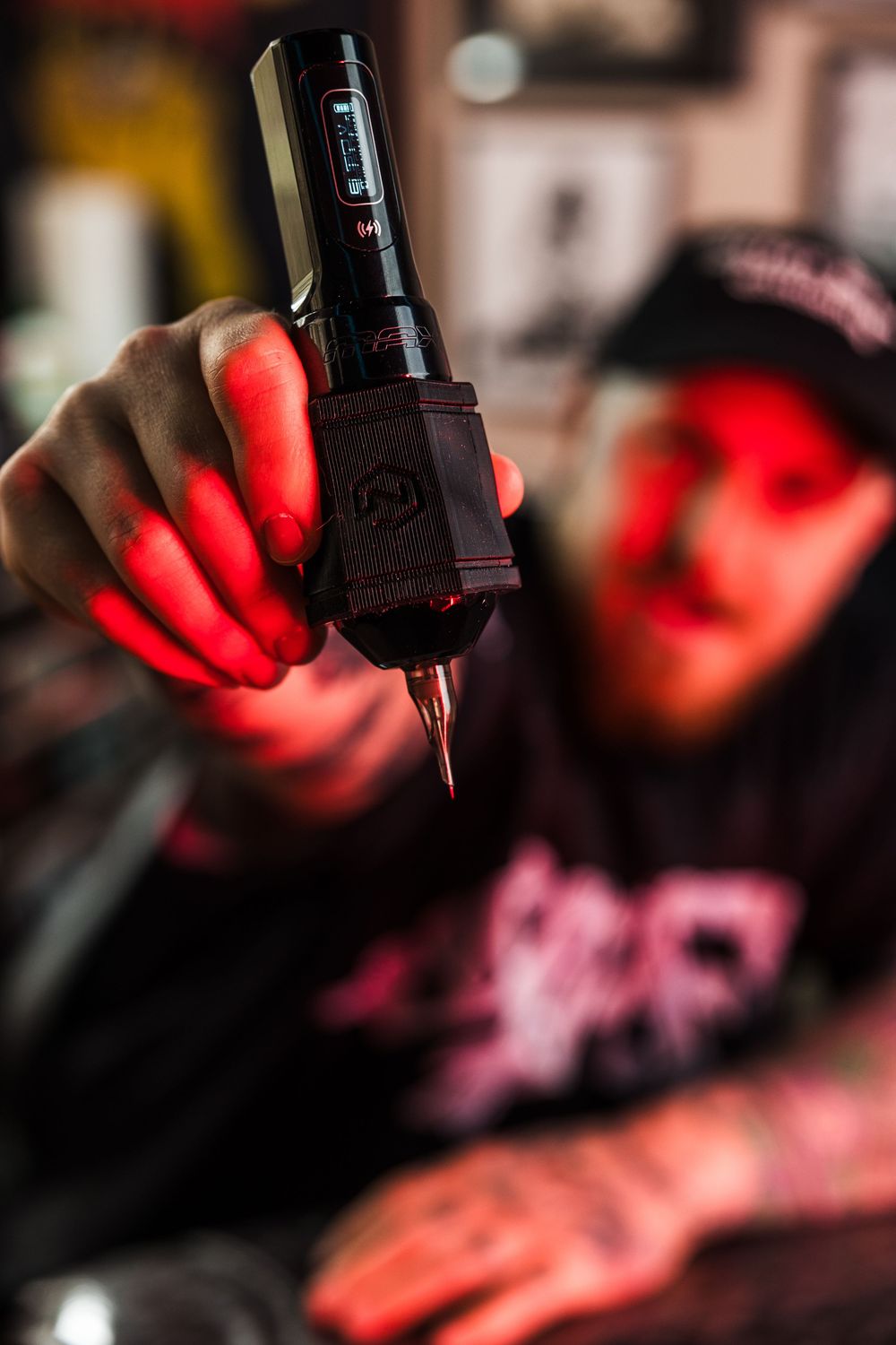 Tattoo artist holding a professional wireless tattoo machine toward camera in red-lit studio.
