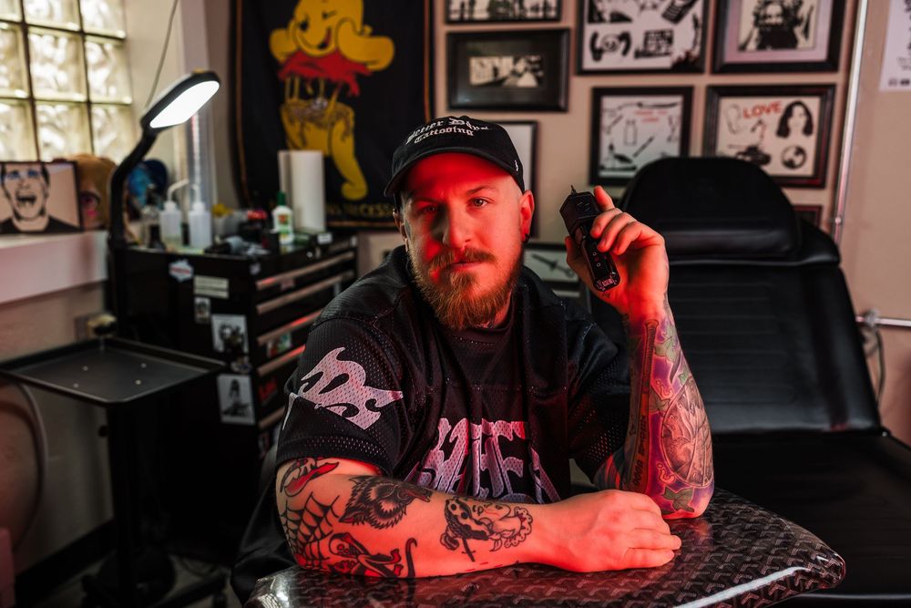 Bearded tattooed man holds tattoo machine while sitting at table in decorated tattoo shop.