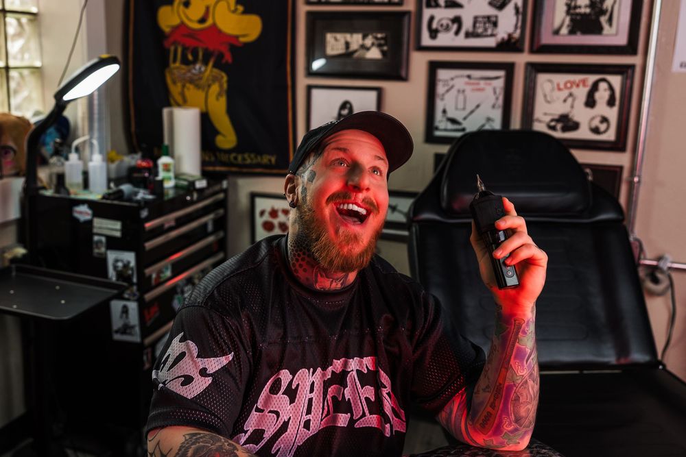 Laughing tattooed man in black hoodie holds tattoo machine in red-lit tattoo studio.