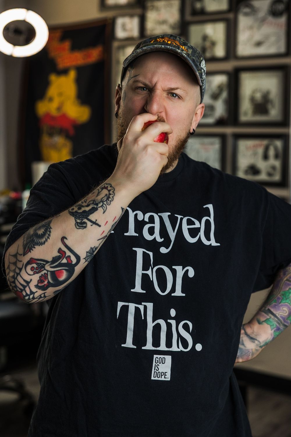 Tattooed man in black Prayed For This God Is Dope tee eating in a tattoo studio.