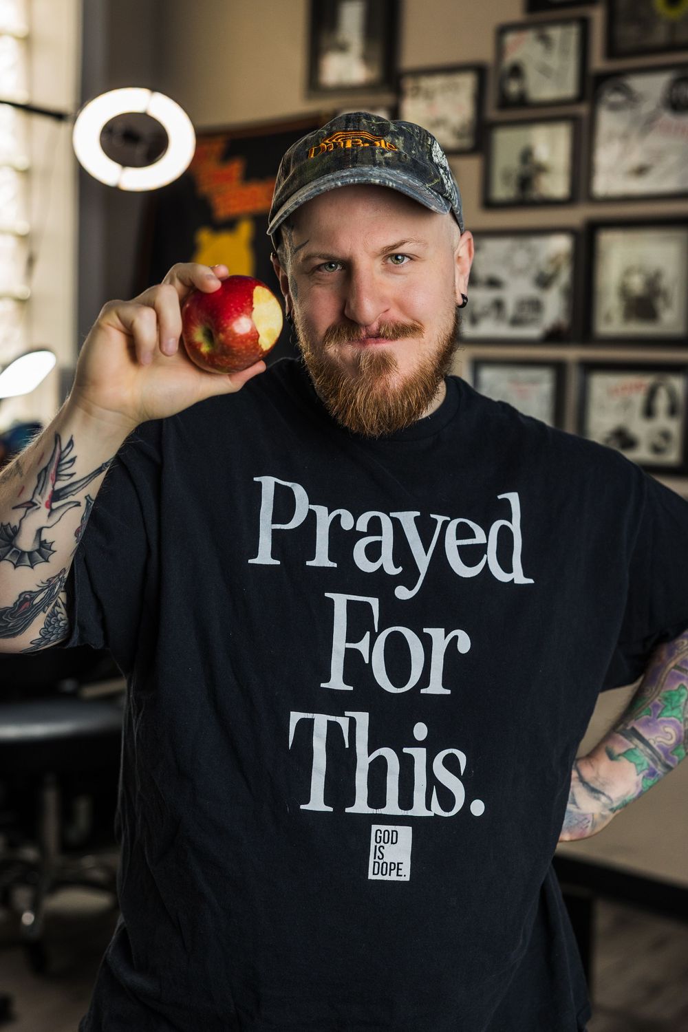 Smiling tattooed man holds red apple wearing Prayed For This black tee in tattoo shop.