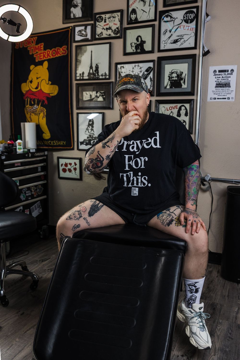 Heavily tattooed man sits on black chair in tattoo studio wearing Prayed For This shirt.
