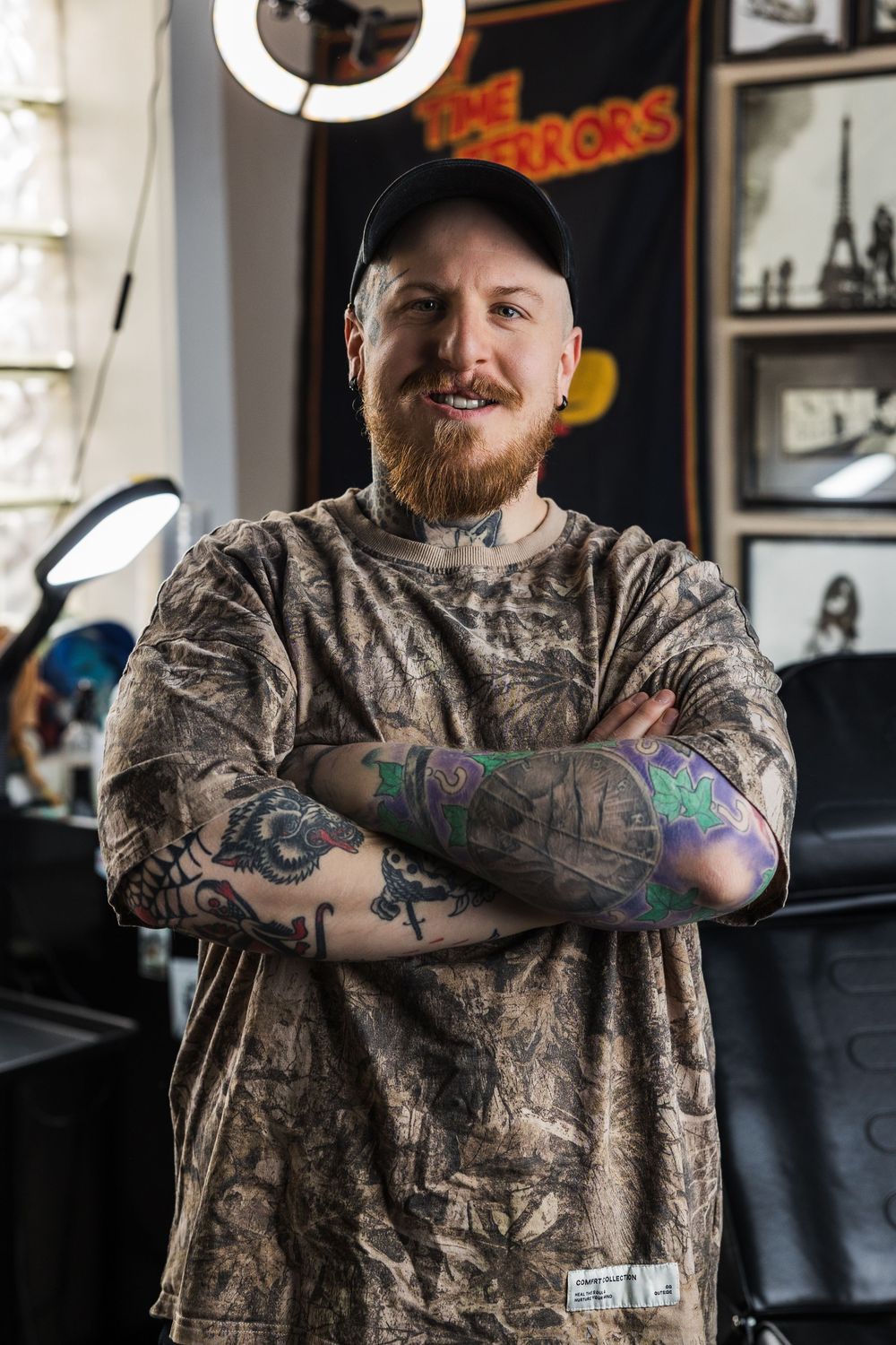 Smiling tattooed bearded man in camo shirt stands with arms crossed in tattoo studio.