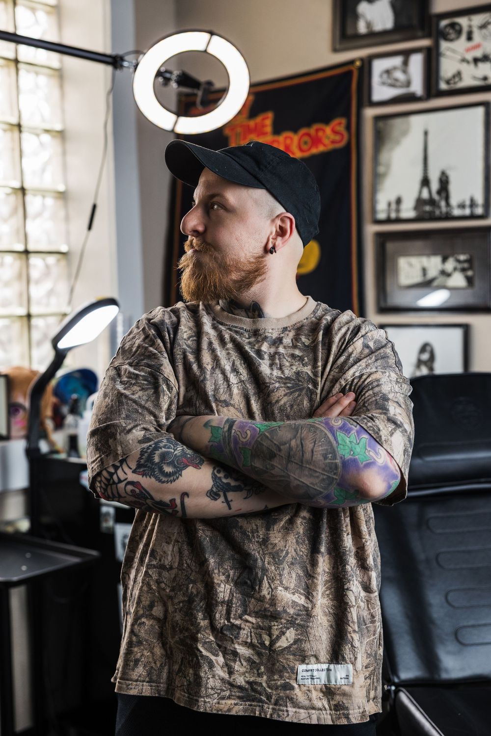 Tattooed bearded man in camo shirt and black cap stands arms crossed in tattoo shop.