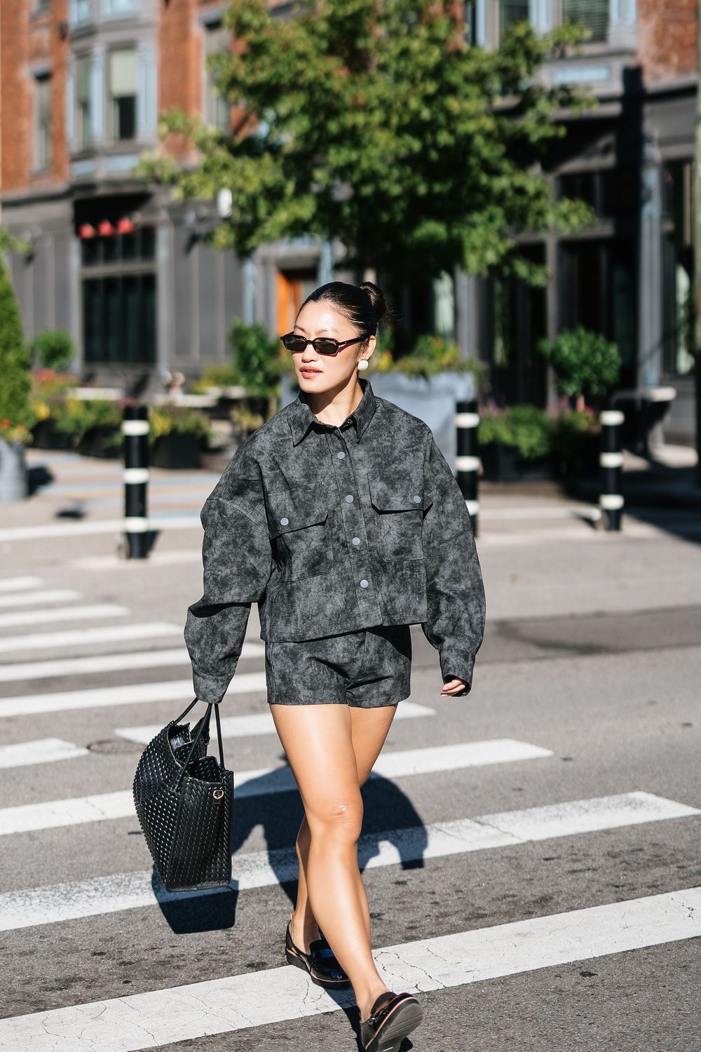 Woman in dark printed jacket and shorts crossing a zebra crossing, carrying a black mesh bag, wearing sunglasses.