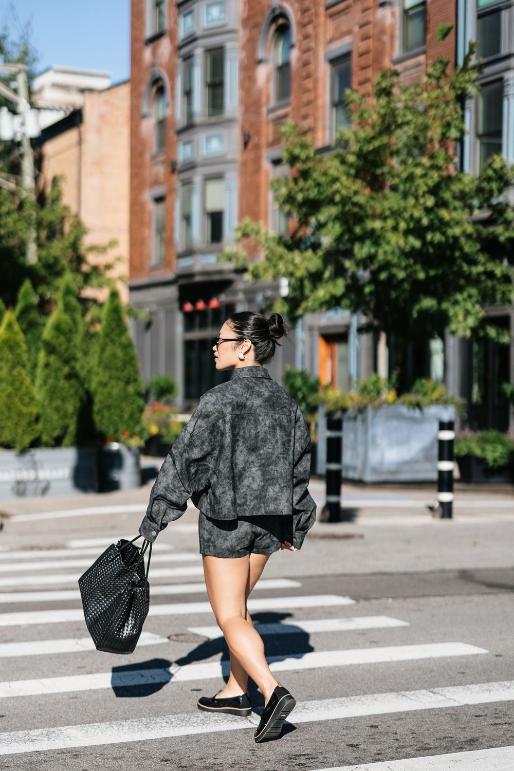 Woman in dark outfit crossing urban street crosswalk, carrying mesh bag, brick buildings and trees in background.