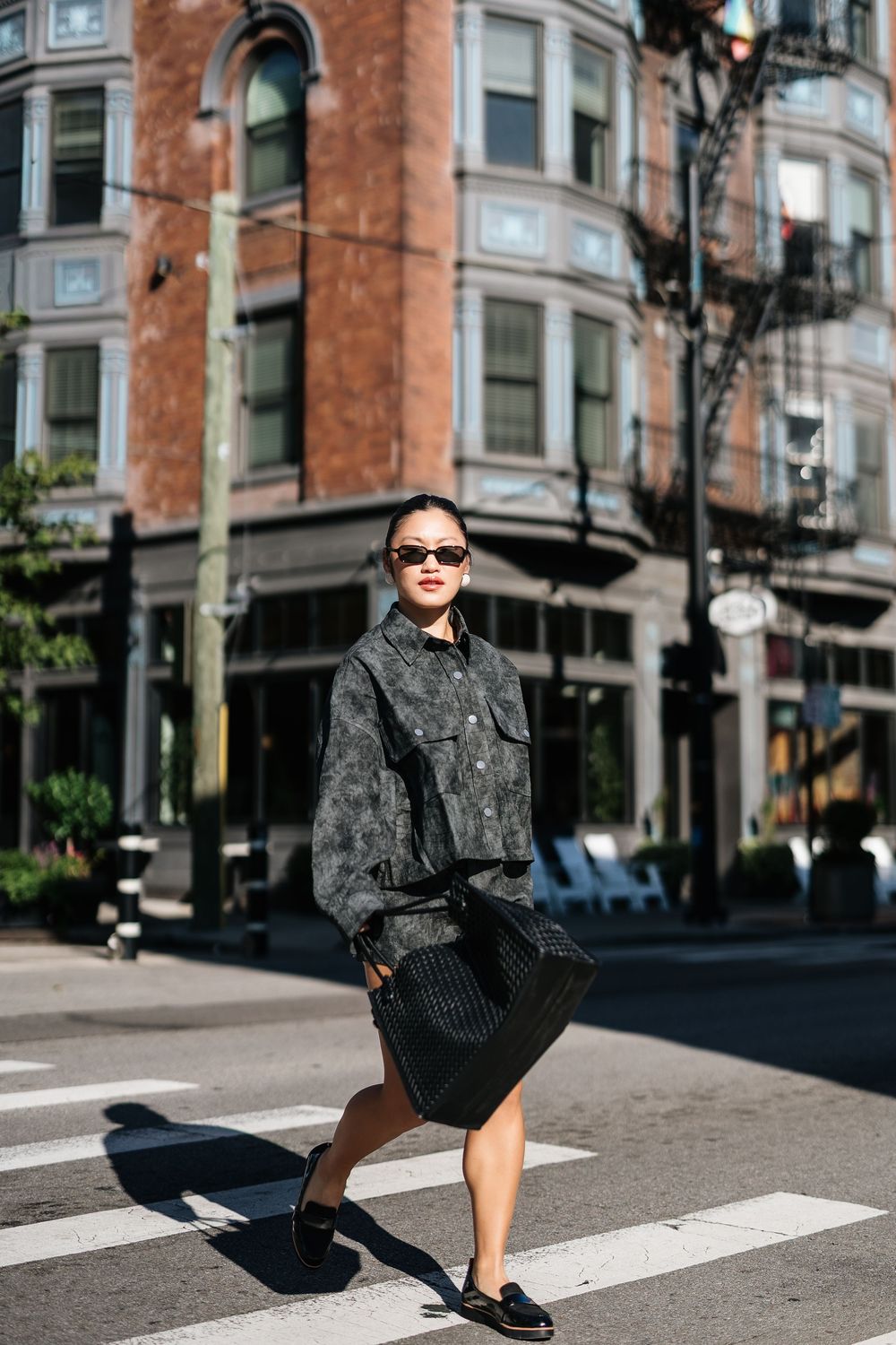 Woman in dark jacket and skirt crossing urban street crosswalk wearing sunglasses and loafers.