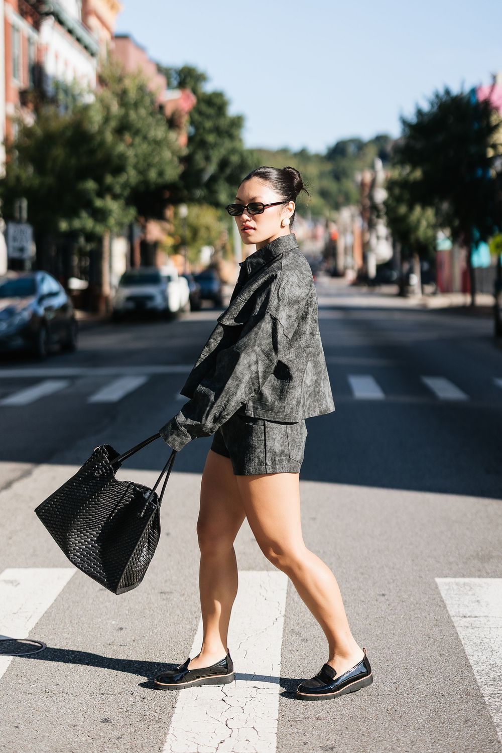 Woman in gray jacket and shorts crossing a street, carrying a black woven tote bag, wearing sunglasses and loafers.