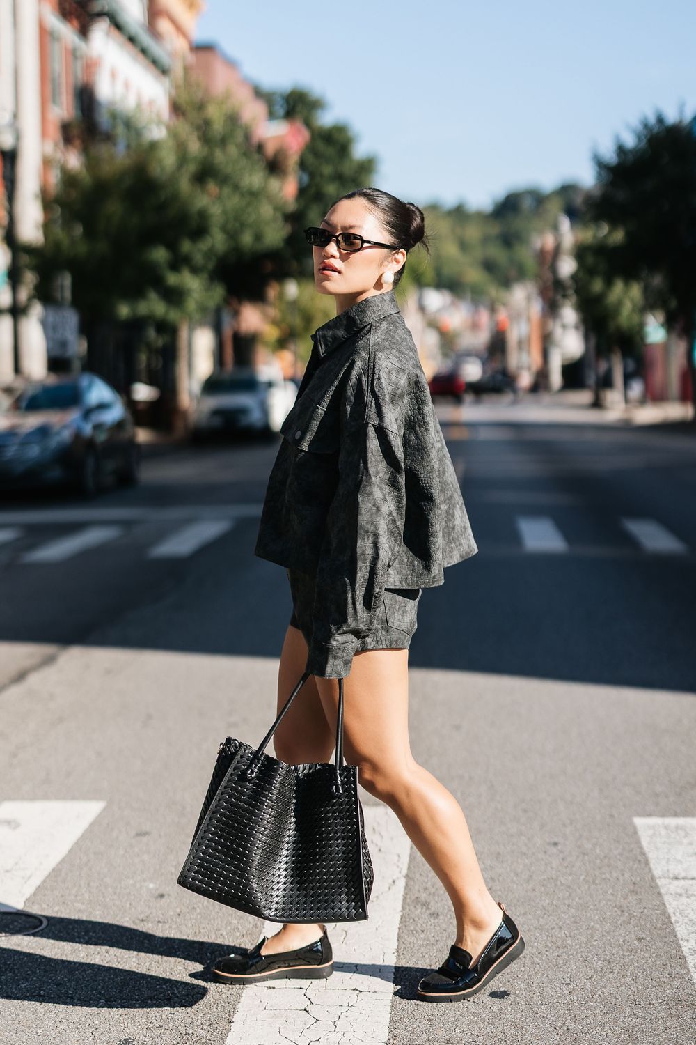 Stylish woman crossing street in dark oversized jacket, shorts, loafers, sunglasses, carrying black woven tote bag.