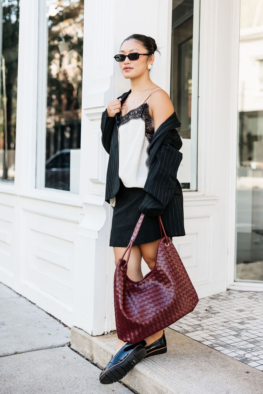 Woman in pinstripe blazer, white lace cami, black mini skirt, loafers, and burgundy woven bag on city sidewalk.