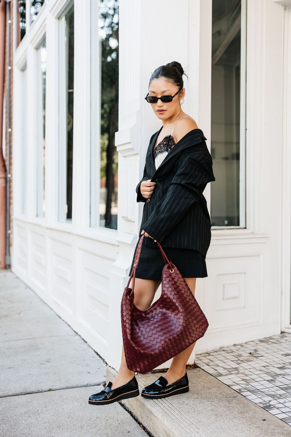 Woman in black off-shoulder dress, striped blazer, loafers, sunglasses, carrying burgundy woven hobo bag on city sidewalk.