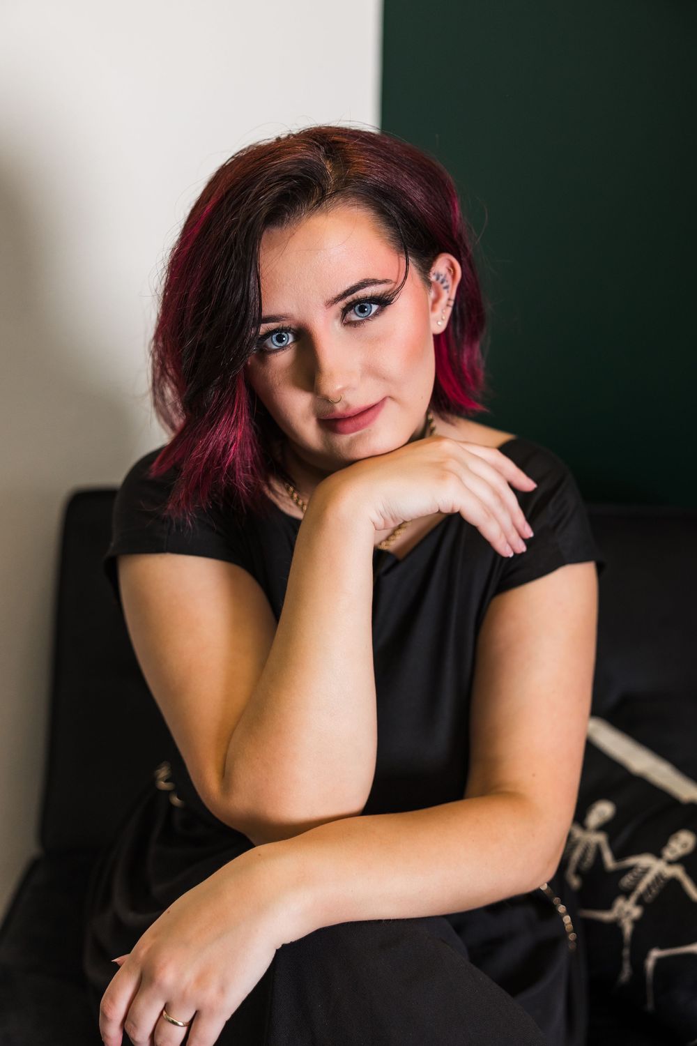 Young woman with pink hair and blue eyes poses confidently in a black dress on a dark sofa.