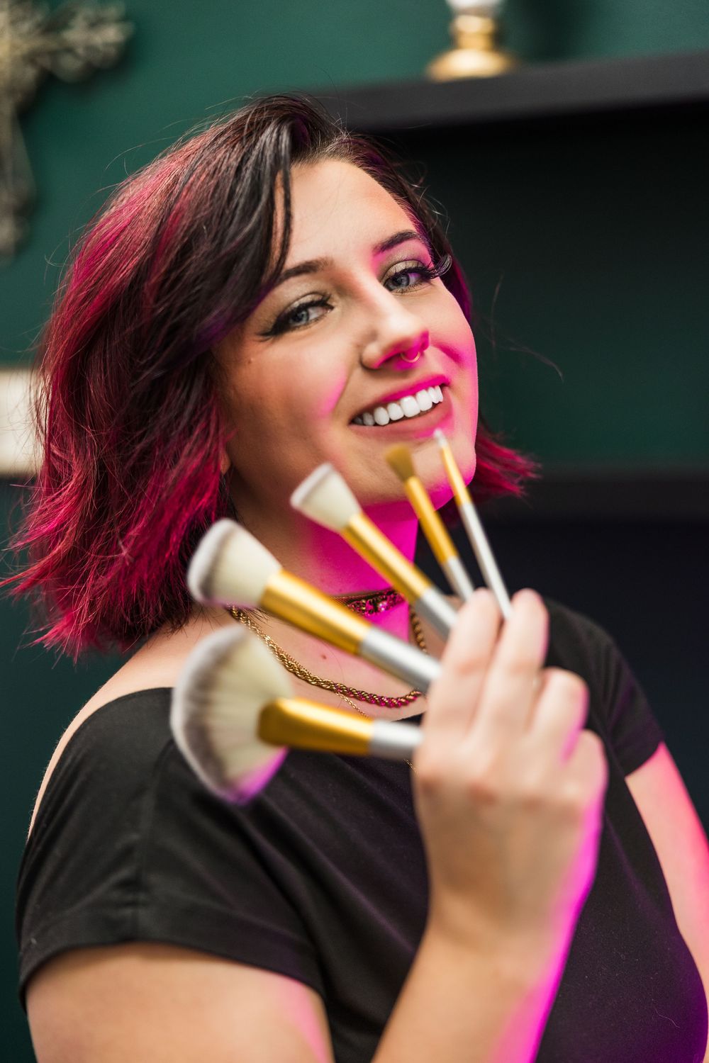 Smiling makeup artist with pink hair holds fan of brushes in a professional beauty studio.