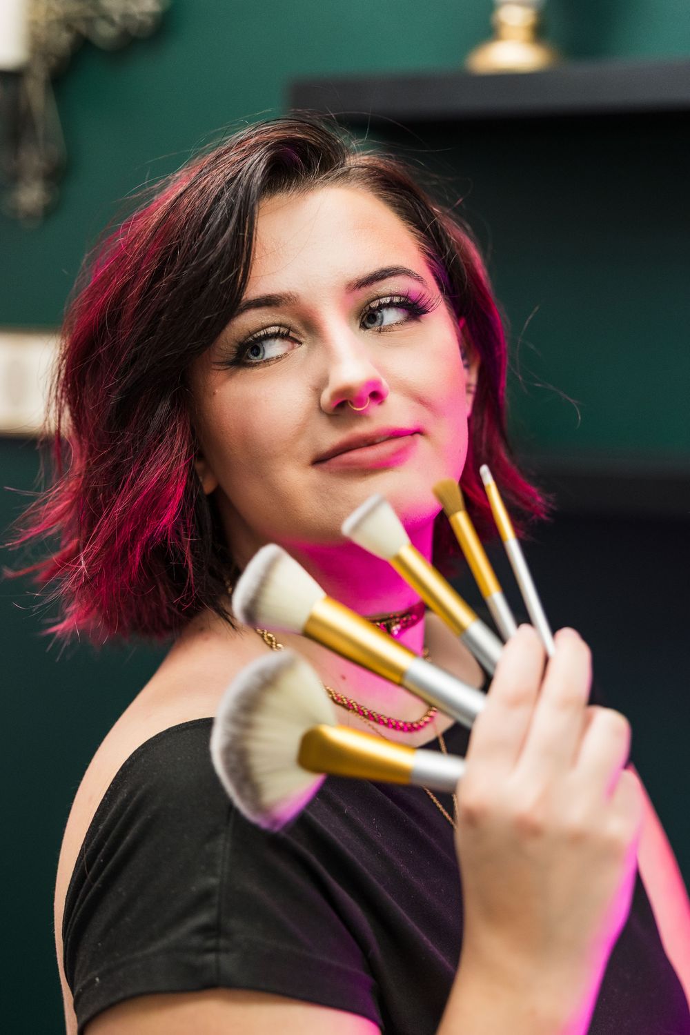 Young woman with pink-highlighted hair holds makeup brushes toward camera in a beauty studio.