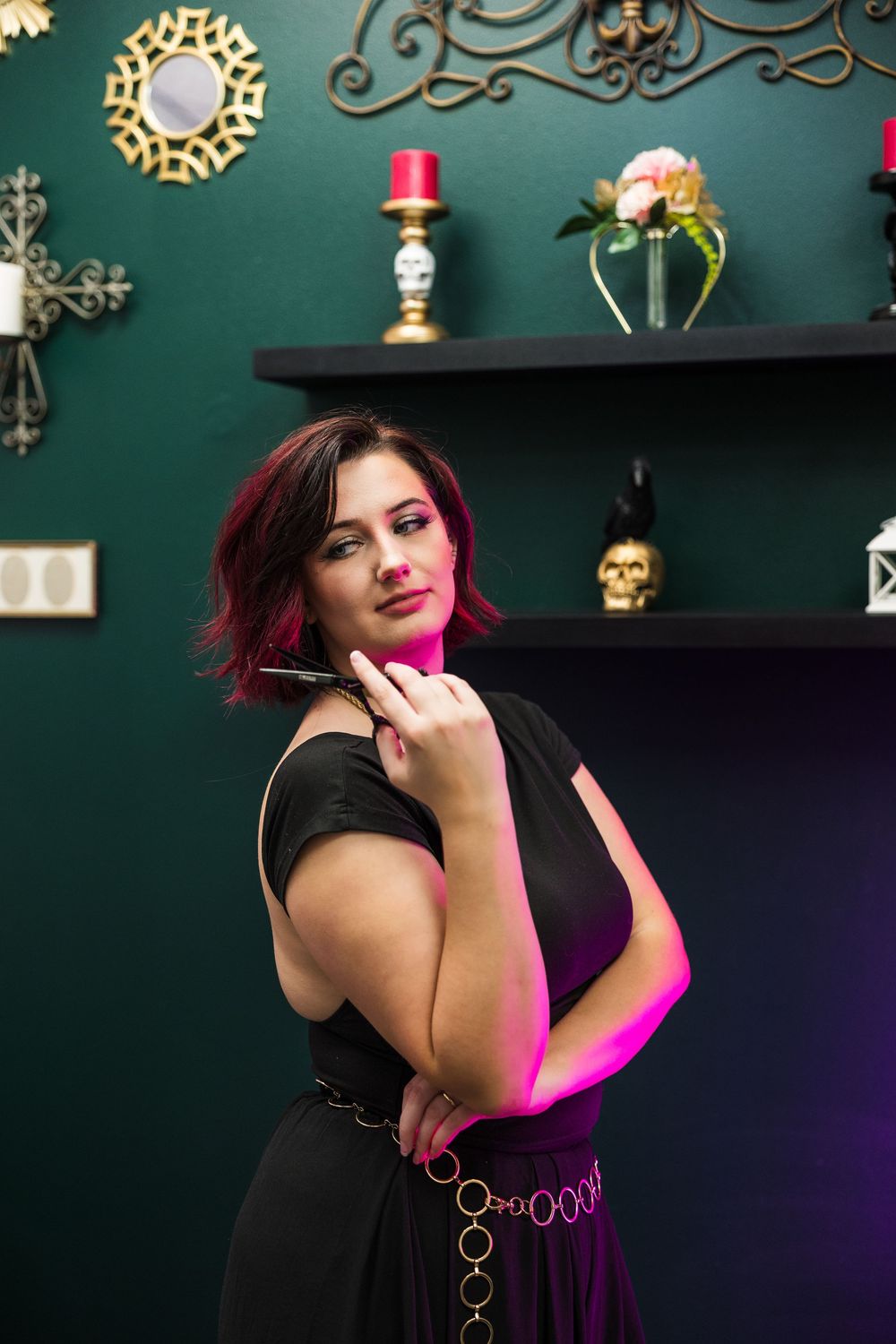 Stylish woman in black dress poses with scissors in a dark green decorated salon.