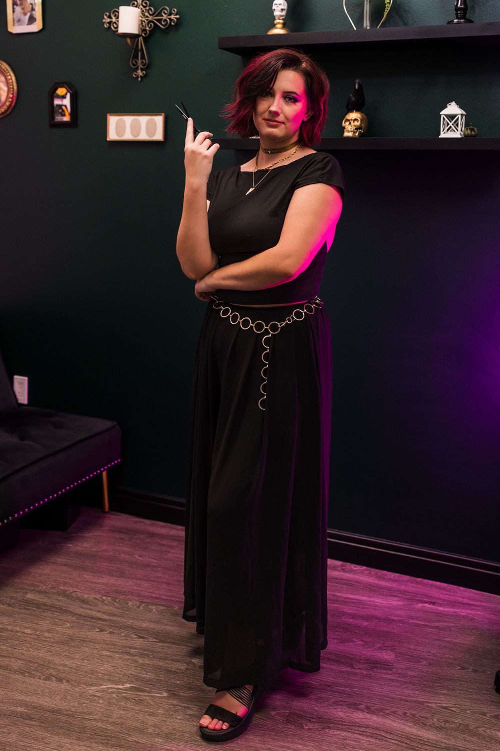 Woman in elegant black maxi dress with gold chain belt poses in a moody green studio.