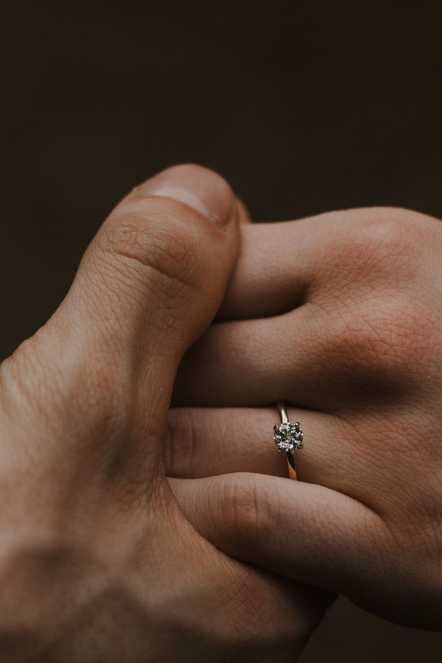 An elegant solitaire diamond engagement ring shown on a hand against a dark background.