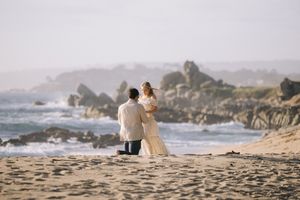 Two people in warm clothing stand together on a rocky beach coastline with crashing waves and misty atmosphere.