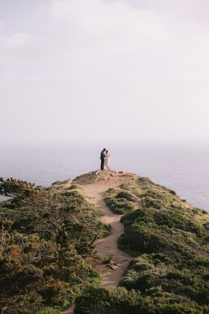 Two distant silhouettes stand together on a dramatic coastal cliff overlooking the misty ocean along a winding dirt trail.