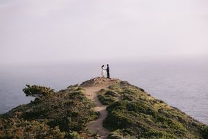 Two silhouettes stand together on a scenic coastal cliff overlooking a misty ocean view along a winding dirt path.