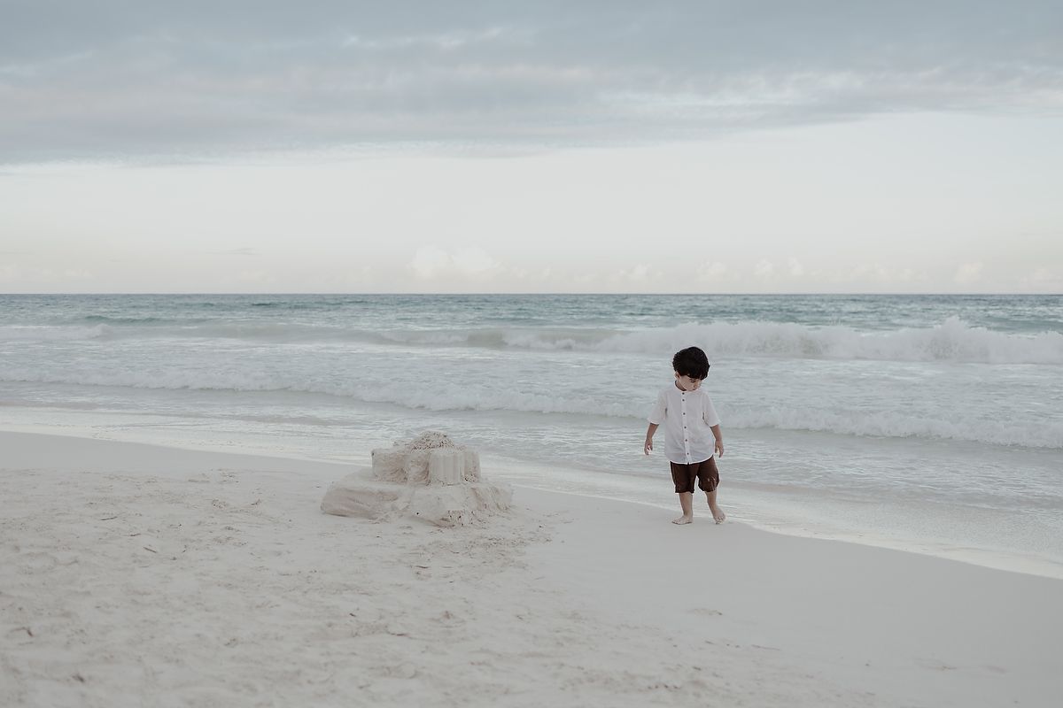 A young child stands on a white sandy beach near a sandcastle, gazing at the calm ocean under a cloudy sky.