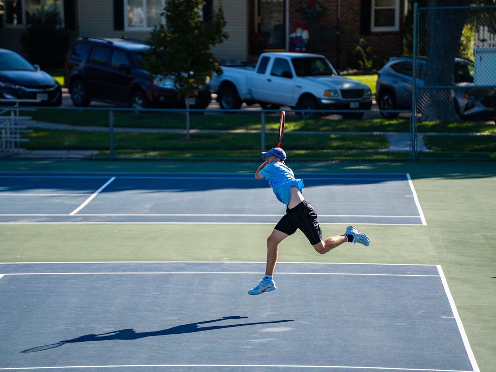 Tennis player in light blue shirt hits a running forehand shot on an outdoor court.