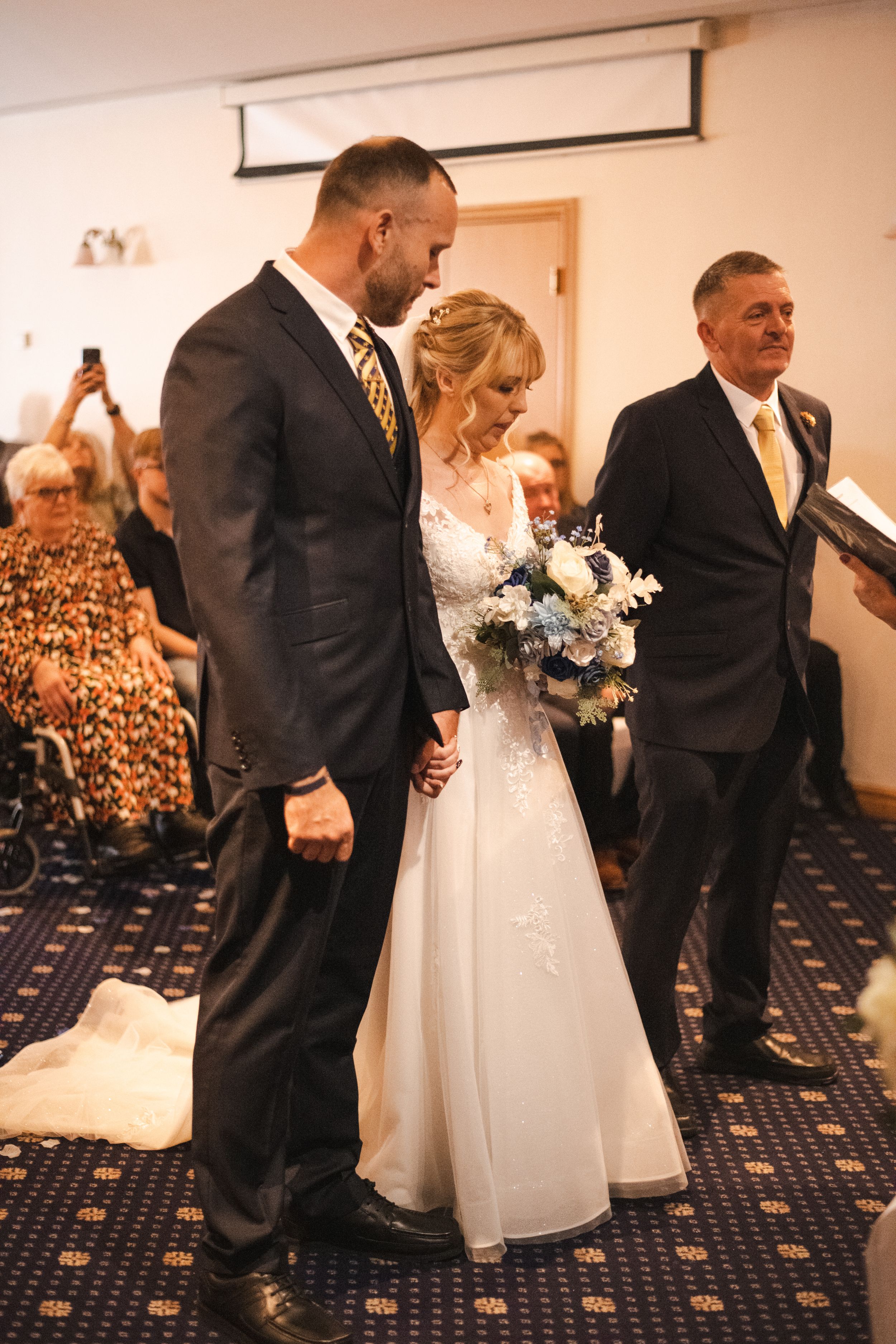 Wedding ceremony in a carpeted room with attendees watching from decorated chairs.