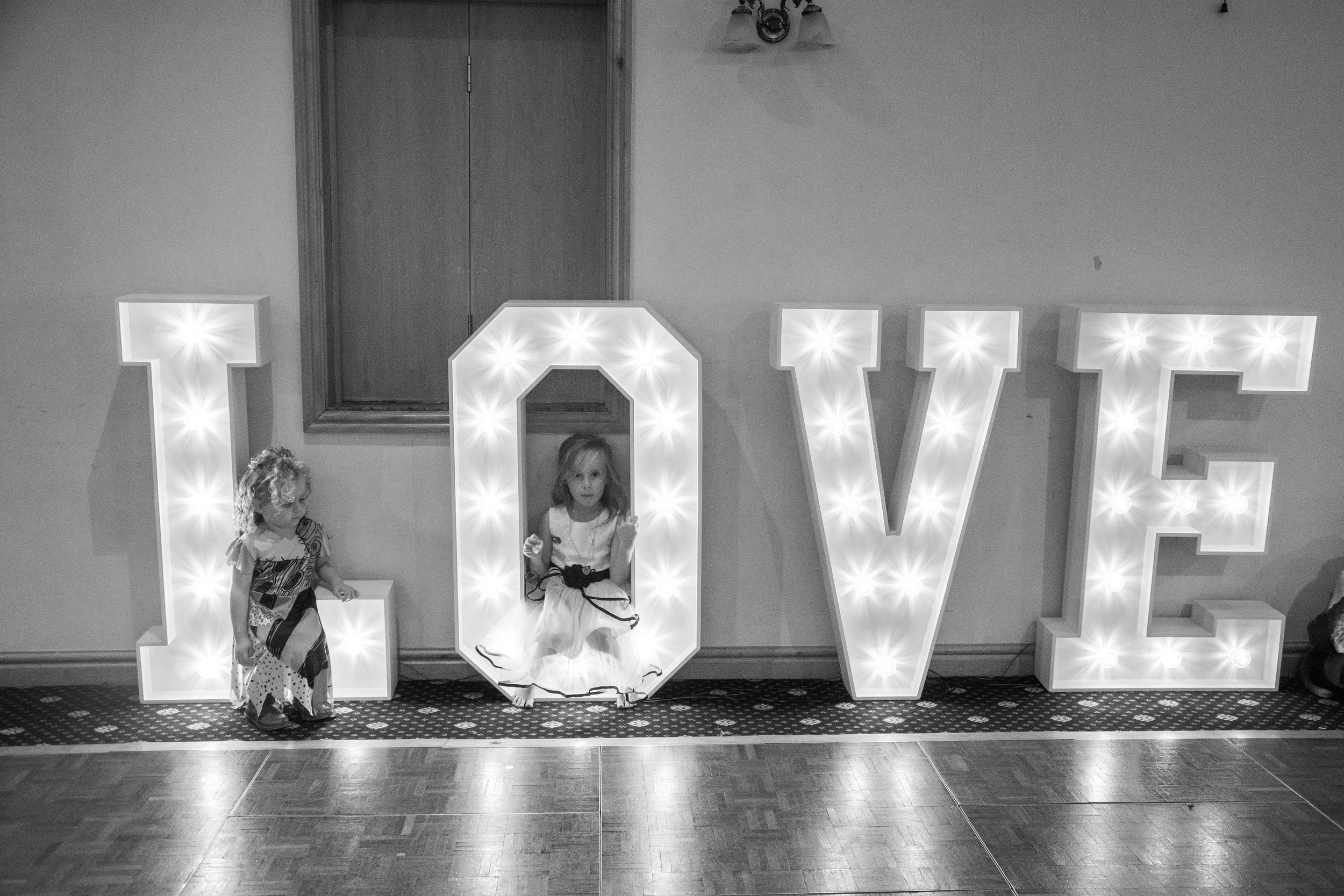 Two children sitting beside illuminated LOVE marquee letters at wedding reception.
