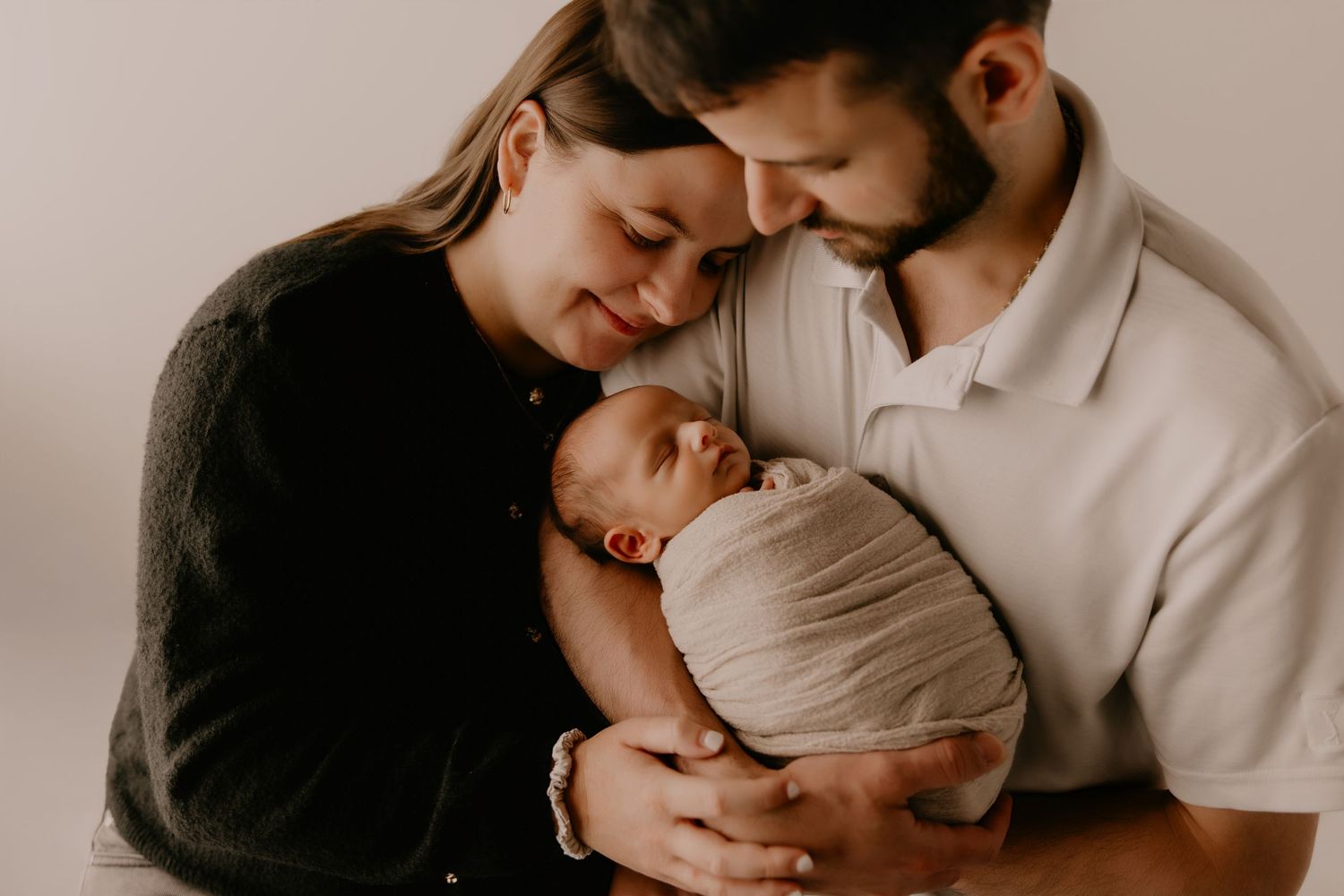 New parents tenderly cradle their sleeping newborn baby wrapped in white, sharing an intimate family moment.