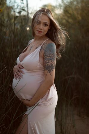 A woman in a light pink dress poses outdoors during golden hour among tall grass and trees.