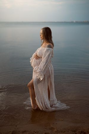 A figure in a white lace dress sits contemplatively at the edge of calm waters during golden hour.
