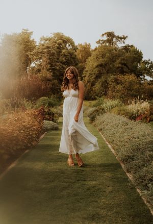 Young woman in flowing white dress walks through lush garden path in golden hour sunlight.