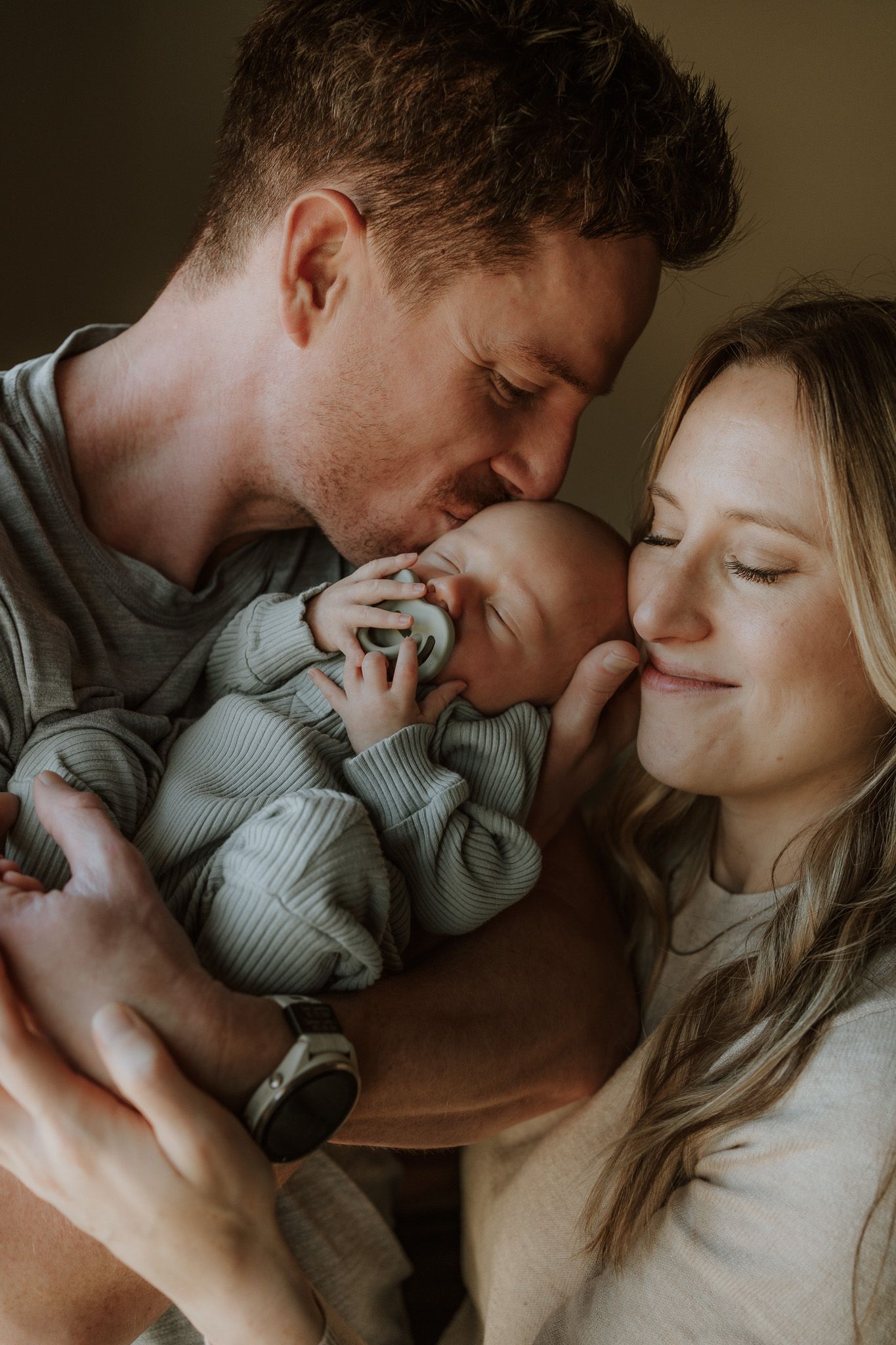 A couple tenderly kisses their newborn baby dressed in a green striped onesie in a warm, intimate portrait.